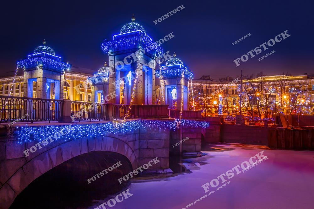 Фотография показывает красочно освещенный мост Ломоносова в Санкт-Петербурге, украшенный праздничной иллюминацией в зимний вечер. Синие огни украшают архитектурные элементы моста, придавая ему волшебный вид, который отражается в темных водах реки ниже. Это изображение идеально подходит для использования в рождественских и новогодних публикациях, для демонстрации красоты зимнего города или в туристических проспектах, рассказывающих о Санкт-Петербурге.